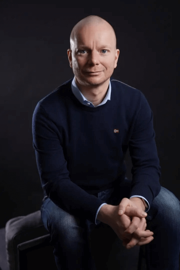 René Fischer sitzt in einem dunkelblauen Pullover auf einem Hocker vor einem schwarzen Hintergrund.