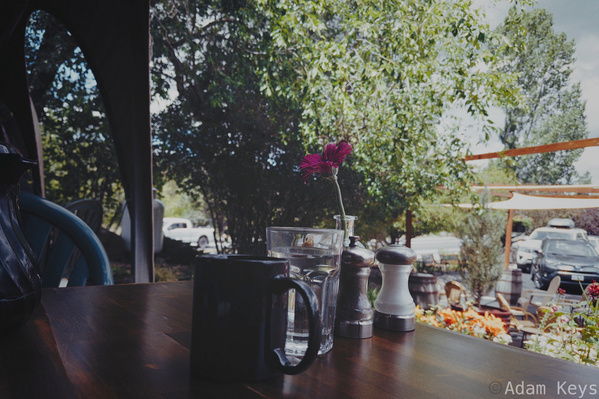A wooden table with a dark mug of coffee, a glass of water, and a single pink flower in a small vase is set near a bright outdoor area with trees and parked cars.