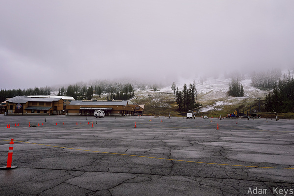 A large empty parking lot with orange traffic cones is situated in front of buildings at the base of a fog-covered mountain with sparse patches of snow and trees.