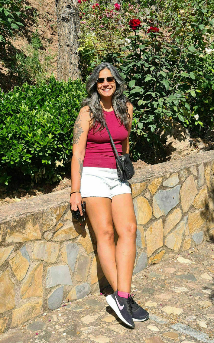 A Brazilian woman wearing a burgundy tank top, white shorts, and sneakers stands smiling beside a stone wall covered in burgundy roses.