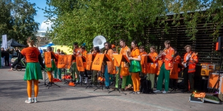 A group of musicians dressed in orange jackets and green attire playing various instruments outdoors. They are accompanied by a conductor facing them, wearing a green skirt and orange tights. The setting is a daytime event with trees and a few tents visible in the background.