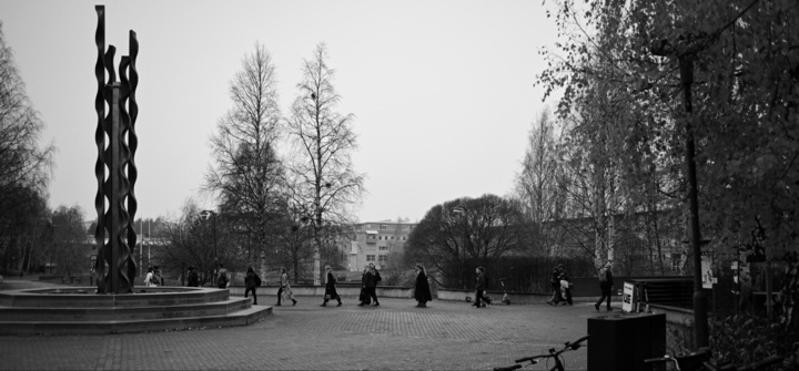 A black and white image of a park scene with people walking. In the foreground, there is a tall, abstract sculpture made of intertwined, wavy metal bars. Bare trees and some buildings are visible in the background.