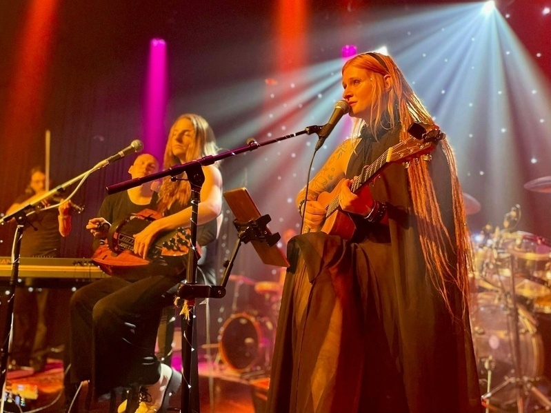 Anna Murphy with a Hurdy Gurdy and Mariana Semkina with an acoustic guitar, sitting on chairs, singing in a microphone, on a stage with coloured lights.