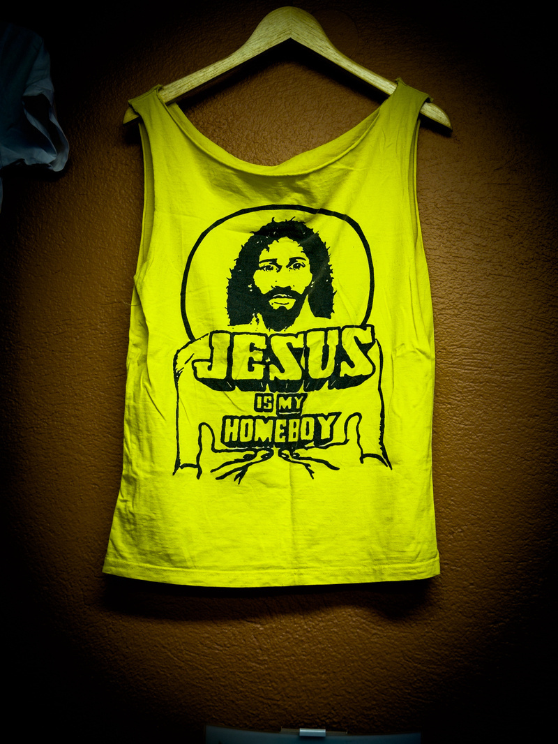  A yellow tank top with the text &ldquo;JESUS IS MY HOMEBOY&rdquo; and an illustration of Jesus hangs on a wooden hanger against a brown textured wall.