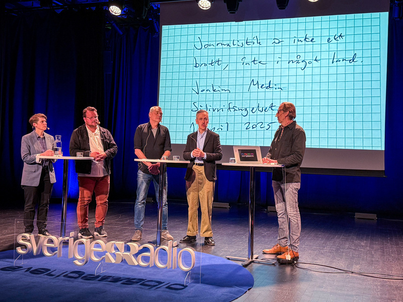 Auto-generated description: Five people are standing on a stage next to tall tables, with writing on a screen behind them displaying handwritten text.