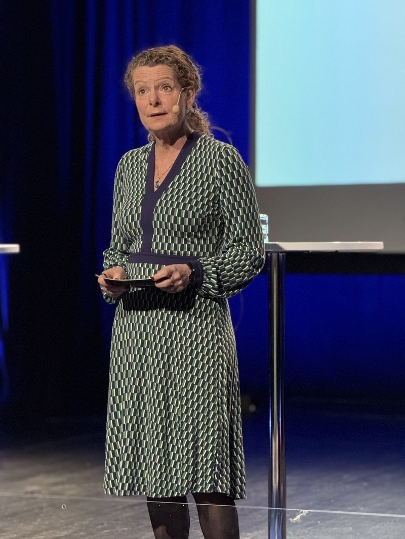 Auto-generated description: A woman in a patterned dress is standing and holding a tablet on a stage.
