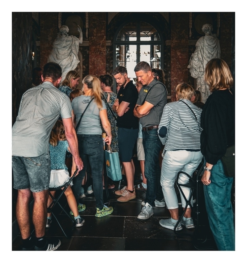 A group of people, some sitting on stools, are closely gathered in an indoor setting that appears to be a museum or historical site with statues in the background.
