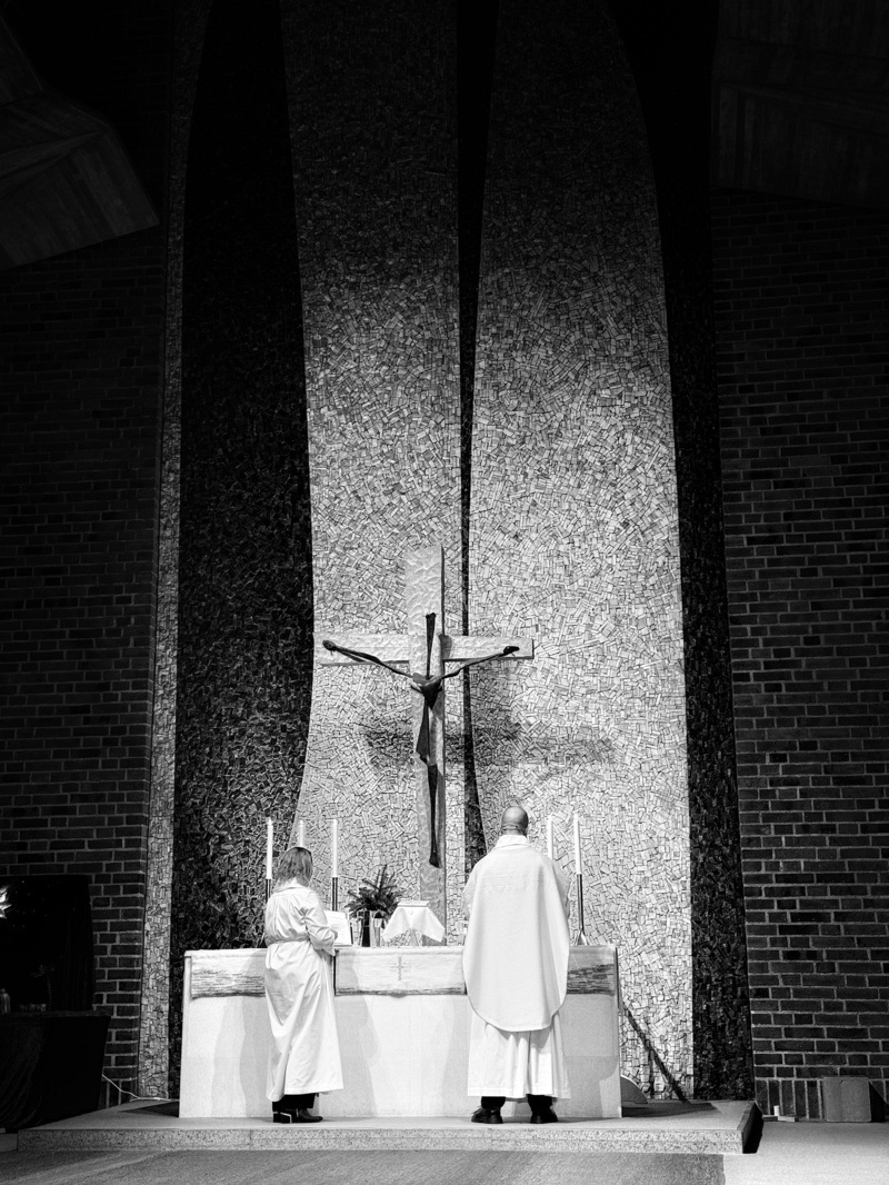 Two people in religious attire stand at an altar in a church. A large crucifix is mounted on a textured wall behind them. The scene is in black and white.