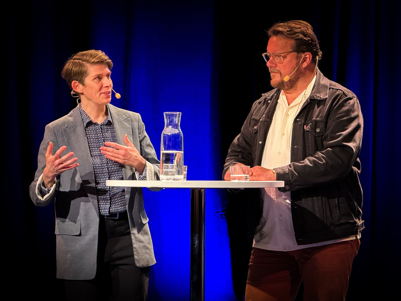 Auto-generated description: Two people are having a discussion on stage at a table with microphones and a water jug, against a blue backdrop.