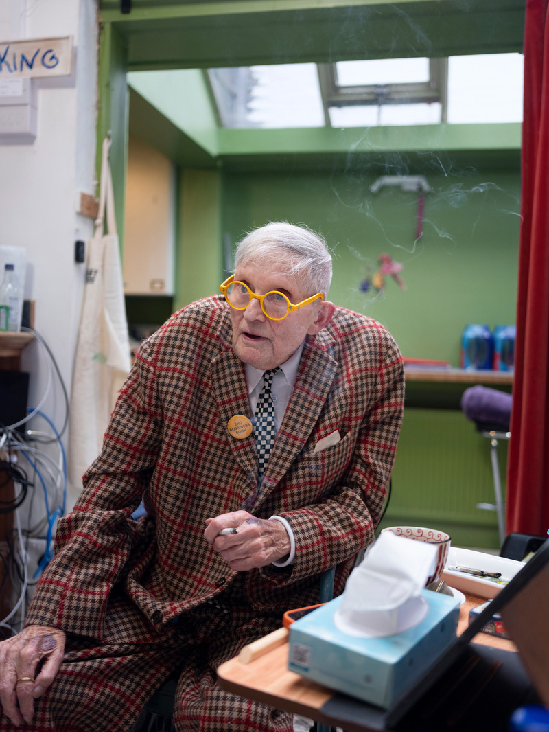 An elderly man in a loud checked suit and round yellow glasses sits forward on a stool in a green studio, cigarette smoke curling past his ear.