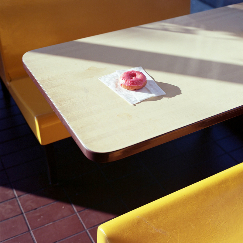 A single pink-frosted donut sits on a paper napkin in the middle of an empty sunlit table, yellow benches on either side.