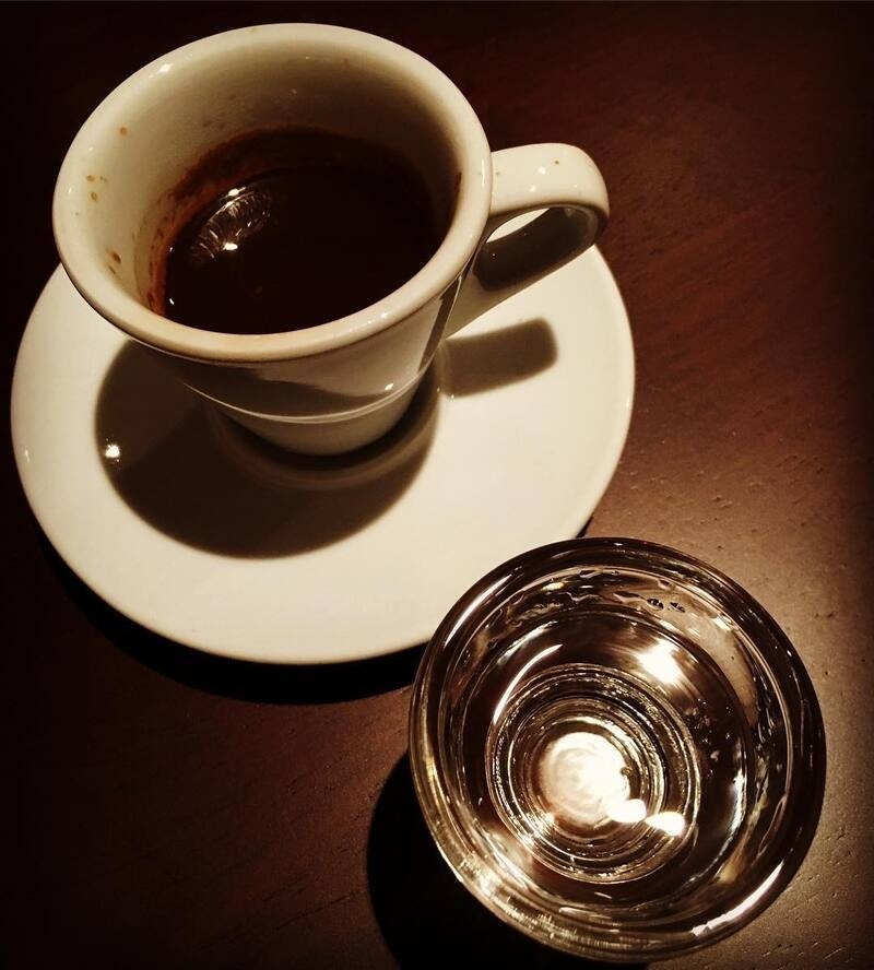 A cup of espresso sits on a white saucer next to a glass of water on a dark wooden table.