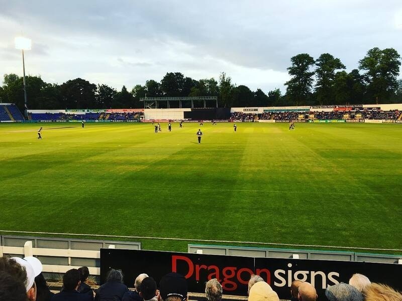 A cricket match is taking place on a well-lit field with spectators watching from the stands.