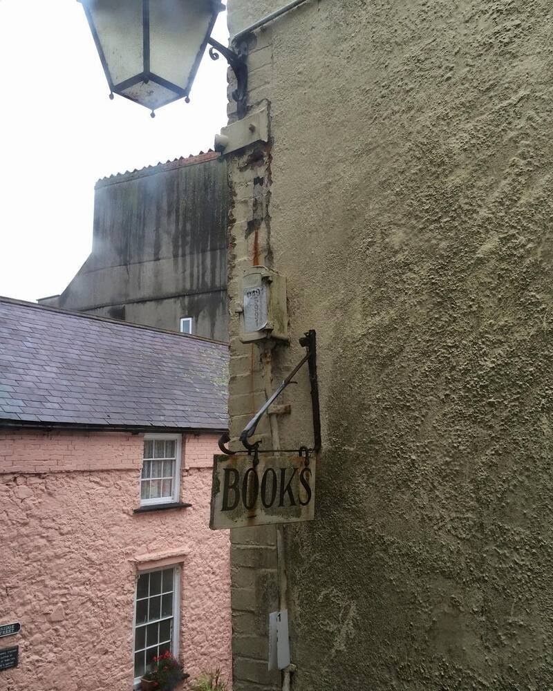 An old-fashioned sign reading BOOKS hangs on a weathered stone wall beneath a vintage street lamp in a quaint, narrow alleyway.