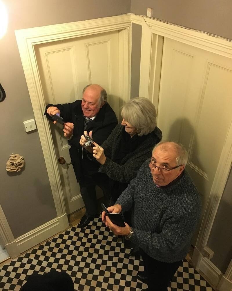 Three older adults are standing closely together in a small corner while holding and looking at their smartphones.