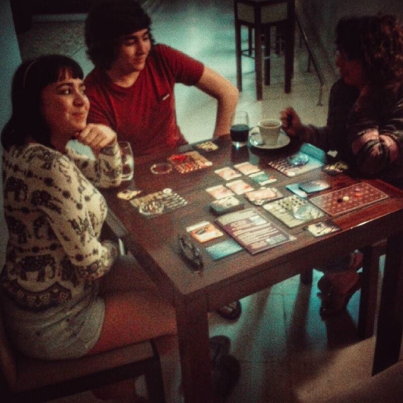Three people are sitting around a table playing a board game, with drinks placed nearby.