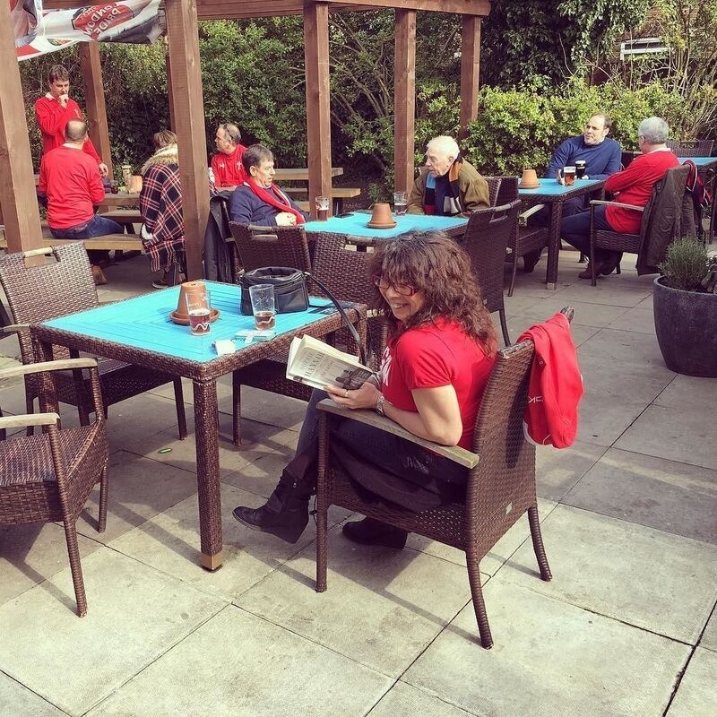 A group of people are sitting and chatting at outdoor tables with blue tablecloths, while a woman in a red shirt reads a book and smiles at the camera.