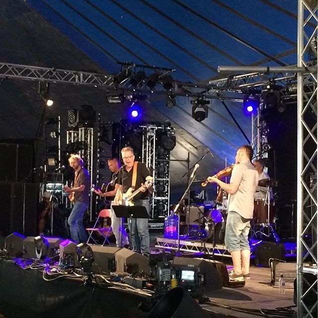 A band is performing on stage under a blue canopy, with musicians playing guitars, violin, and drums amidst stage lighting and sound equipment.