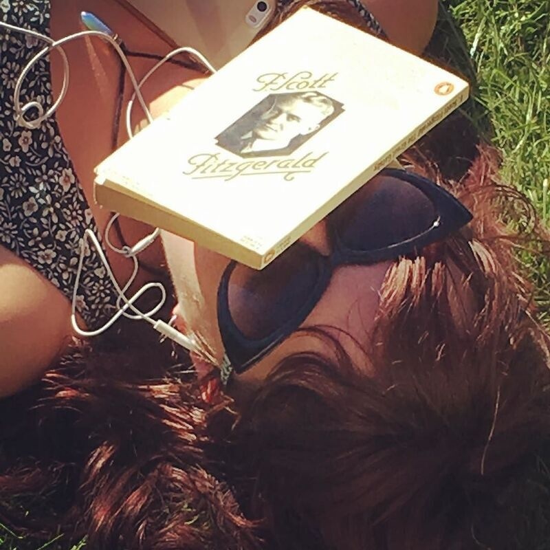 A person wearing sunglasses is lying on the grass with a book by F. Scott Fitzgerald resting on their face and earphones nearby.