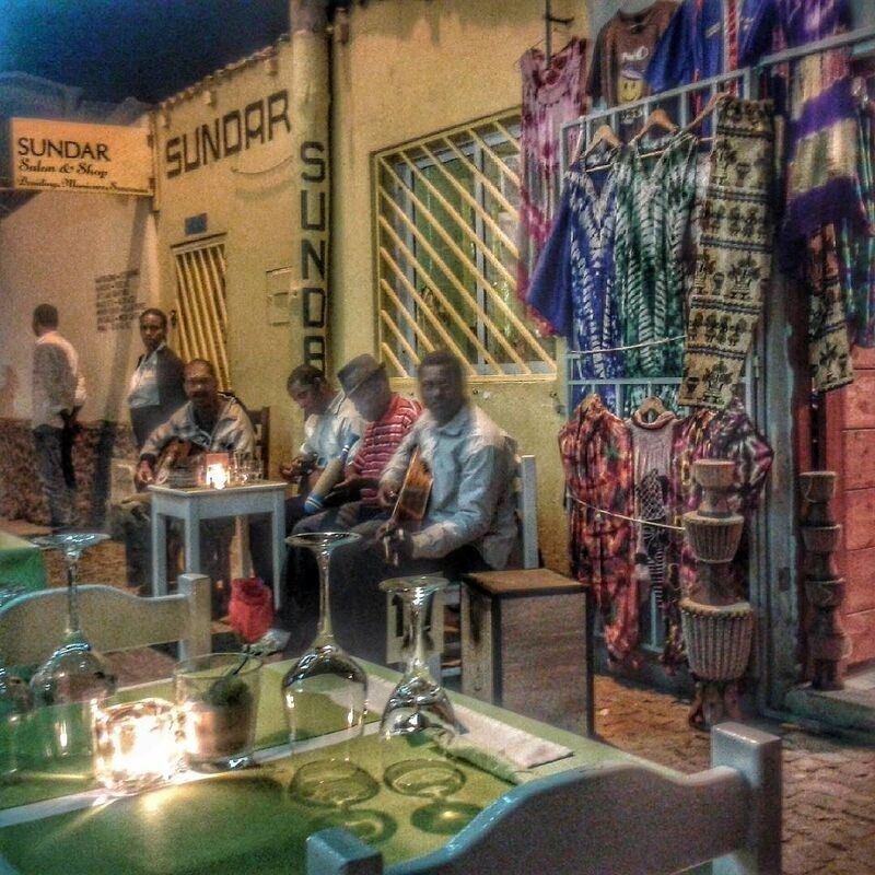 A group of musicians is performing outside a shop named Sundar, while colorful garments are displayed on hangers nearby and tables are set with glasses and candles in the foreground.