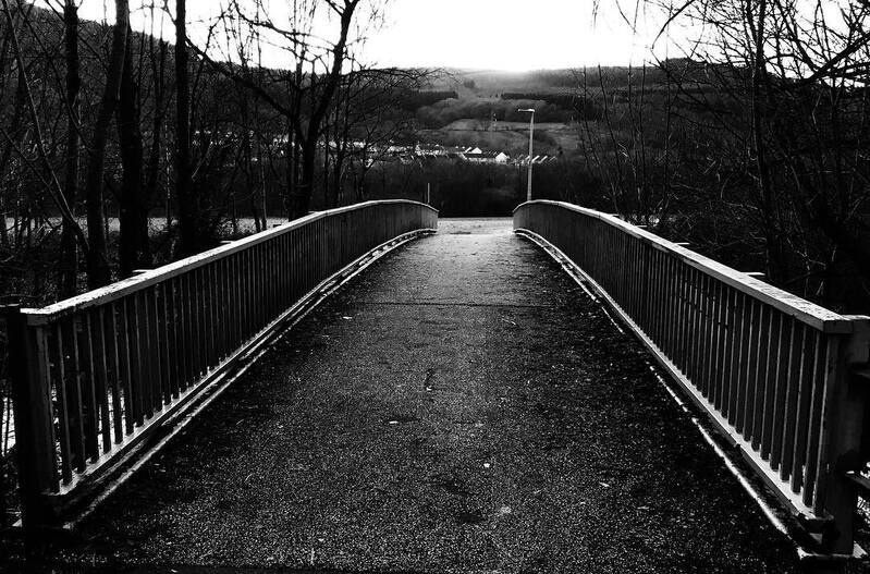 A black and white photo of a bridge with railings on both sides, surrounded by trees and hills in the background.