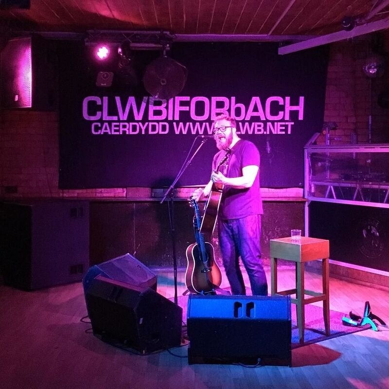 A musician is performing with an acoustic guitar on a small stage under purple lighting at Clwb Ifor Bach in Cardiff.