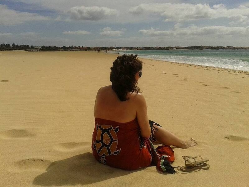 A person wearing a red dress with a turtle design is sitting on the sandy beach, facing the ocean.