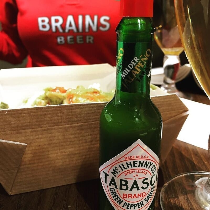 A bottle of Tabasco Green Pepper Sauce is placed on a table with food and drinks, while a person wearing a red BRAINS BEER shirt is in the background.