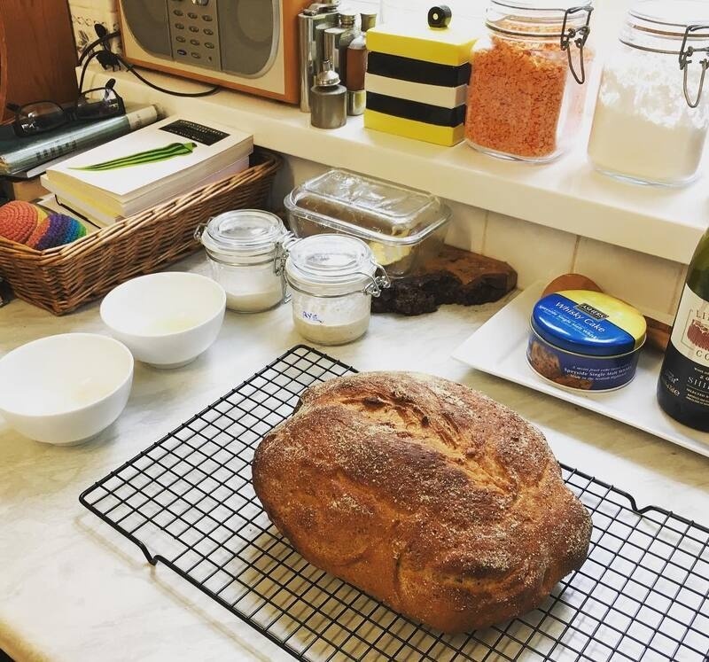 A freshly baked loaf of bread rests on a cooling rack in a kitchen countertop surrounded by various jars, bowls, and kitchen items.