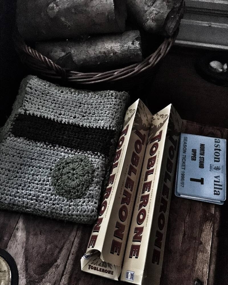A woven fabric piece, three Toblerone chocolate bars, and an Aston Villa season ticket for the 1986/87 season are arranged on a wooden surface.