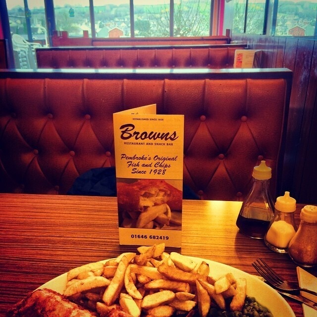 A plate of fish and chips with peas is served on a wooden table beside condiments and a Browns restaurant menu in a booth seating area.