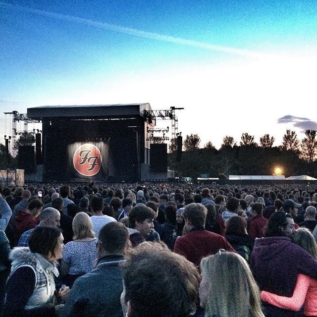 A large crowd gathers outdoors in front of a stage displaying the Foo Fighters logo at dusk.