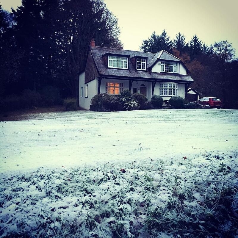 A two-story house with lit windows sits in a snowy yard surrounded by trees, with a red car parked nearby.