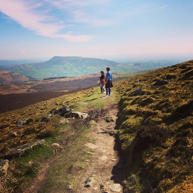 Two people are hiking along a scenic hillside trail with expansive views of green fields and hills in the distance.