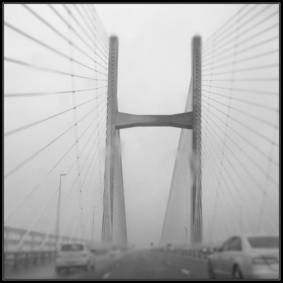 A suspension bridge with two tall towers and multiple cables is shown with vehicles traveling on the road beneath it on a foggy day.