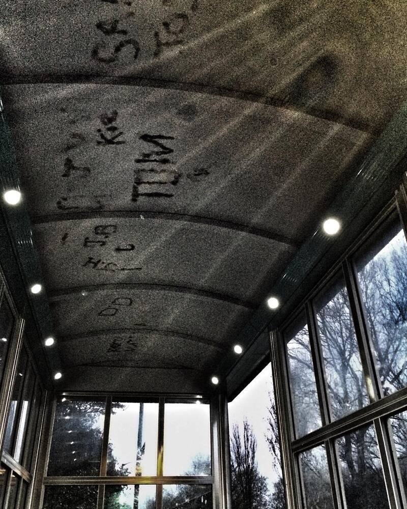 A narrow walkway with large glass windows on the right side and overhead lighting features graffiti on its arched ceiling.