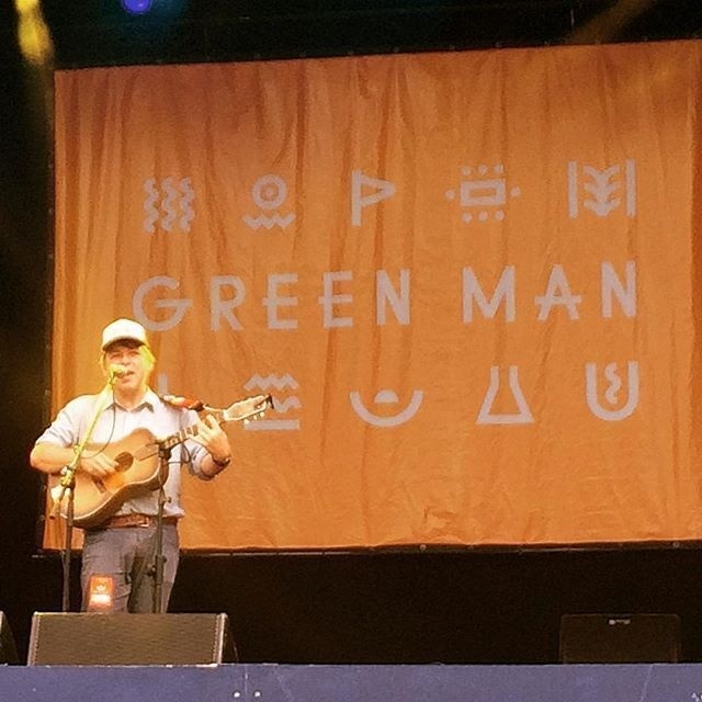 A musician wearing a cap is playing an acoustic guitar and singing on stage in front of a large orange backdrop with the words GREEN MAN and various symbols.