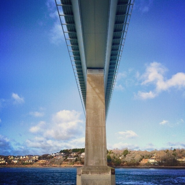 A large bridge is supported by a central concrete pillar, spanning over a body of water with a scenic, tree-lined shoreline and houses in the background.