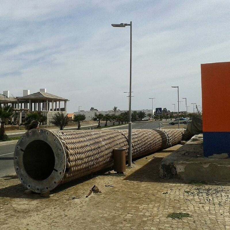 A long, cylindrical metallic object with a textured surface is placed on the ground near a roadside with streetlights and buildings in the background.