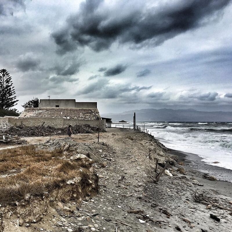 A rocky coastal path with sparse vegetation leads to a building near the shore under a cloudy, stormy sky.
