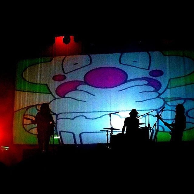 A band is performing on stage with a large cartoon clown face projected as a colorful backdrop behind them.
