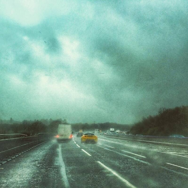 Several vehicles, including a yellow car and a truck, are driving on a wet highway under a gloomy, overcast sky.