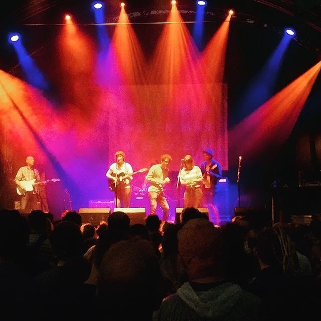A band is performing on stage with colorful spotlights illuminating them while a crowd watches.