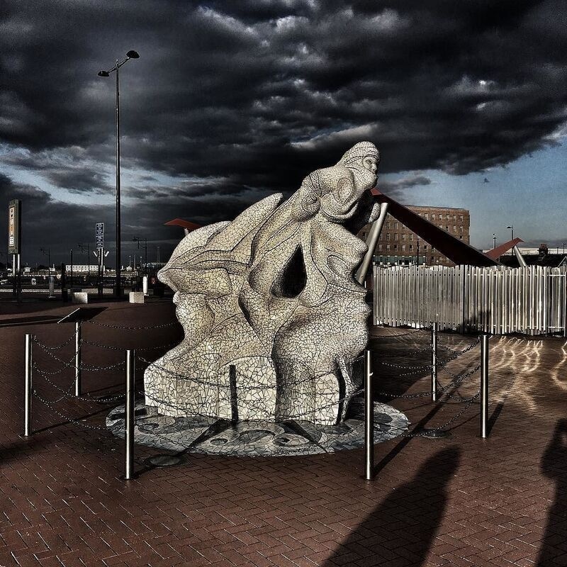A detailed stone sculpture featuring intertwined human figures stands under a dramatic, cloudy sky on a paved area surrounded by a low chain fence.