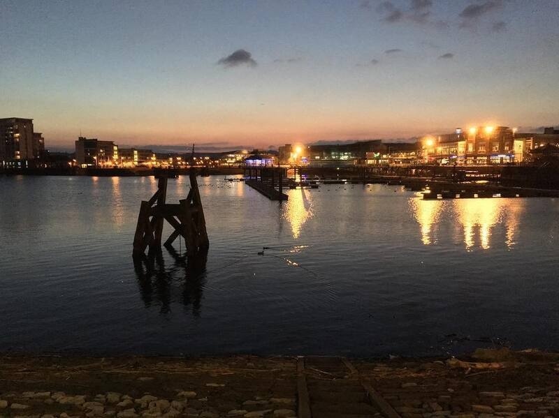 A calm waterfront scene at dusk features illuminated buildings and structures reflecting on the water.