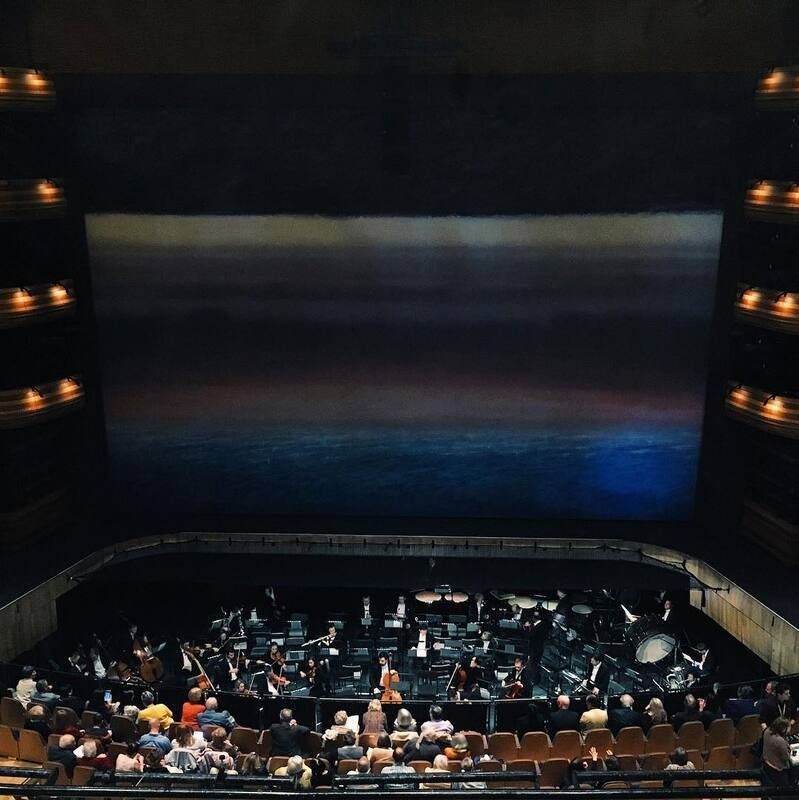 An orchestra is performing on stage in front of an audience inside a dimly lit concert hall.