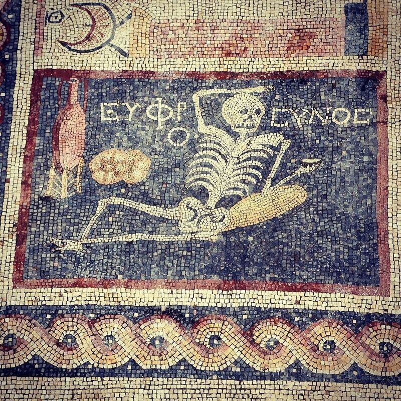 A mosaic depicts a reclining skeleton holding a scroll or tablet, accompanied by Greek text and surrounded by various symbolic objects.