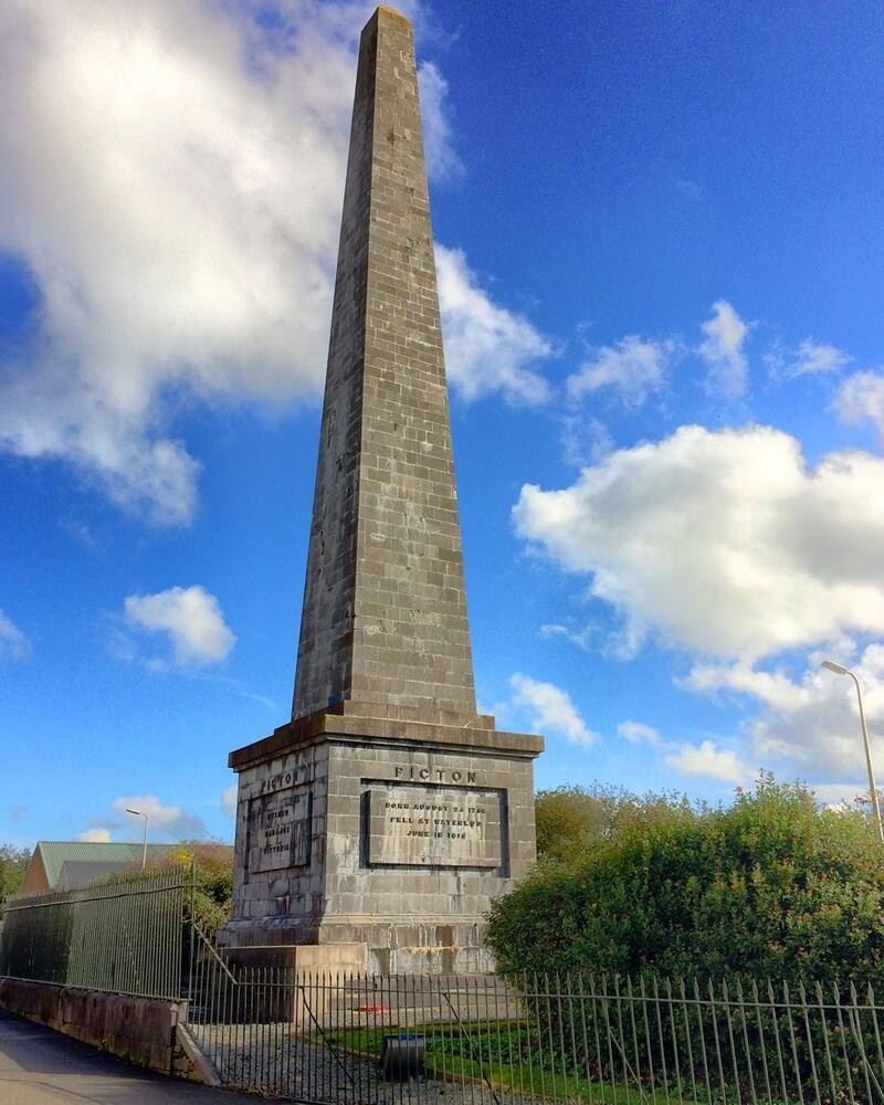 A tall stone obelisk monument stands against a blue sky with scattered clouds, surrounded by greenery and a metal fence.