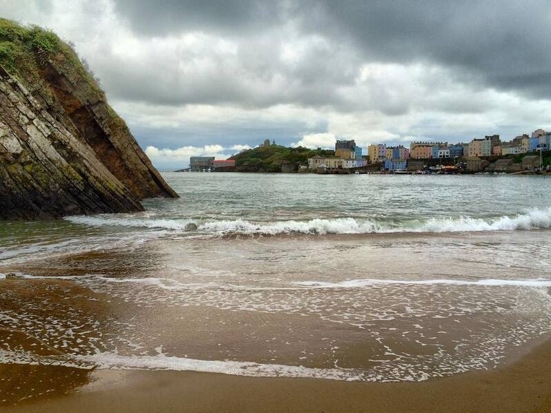 A sandy beach with gentle waves is bordered by rocky cliffs on the left and a town with colorful buildings in the distance under a cloudy sky.