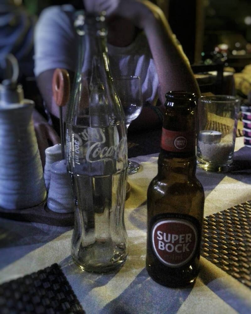 A table is set with an empty Coca-Cola glass bottle, a Super Bock beer bottle, a person in the background, and various dining items in a dimly lit setting.